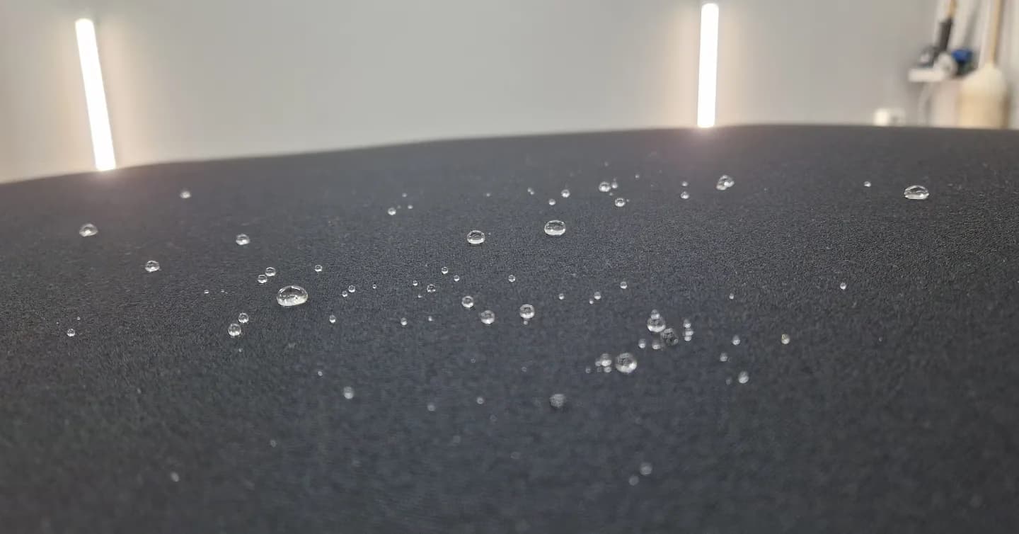 Makroaufnahme von Wasserperlen auf einer frisch versiegelten schwarzen Stoffoberflaeche (Cabrio-Verdeck) unter zwei LED-Leuchtbalken.