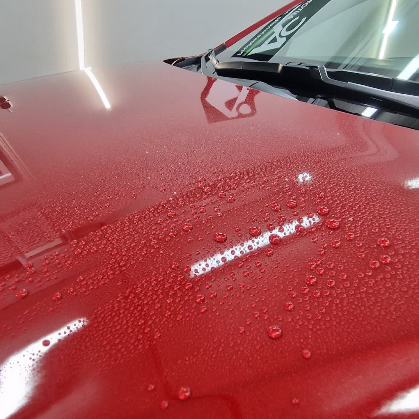 Perlender Wasserfilm auf der roten, keramikversiegelten Motorhaube eines Neuwagens, im Hintergrund die Frontscheibe mit AC-Autopflege-Banner.